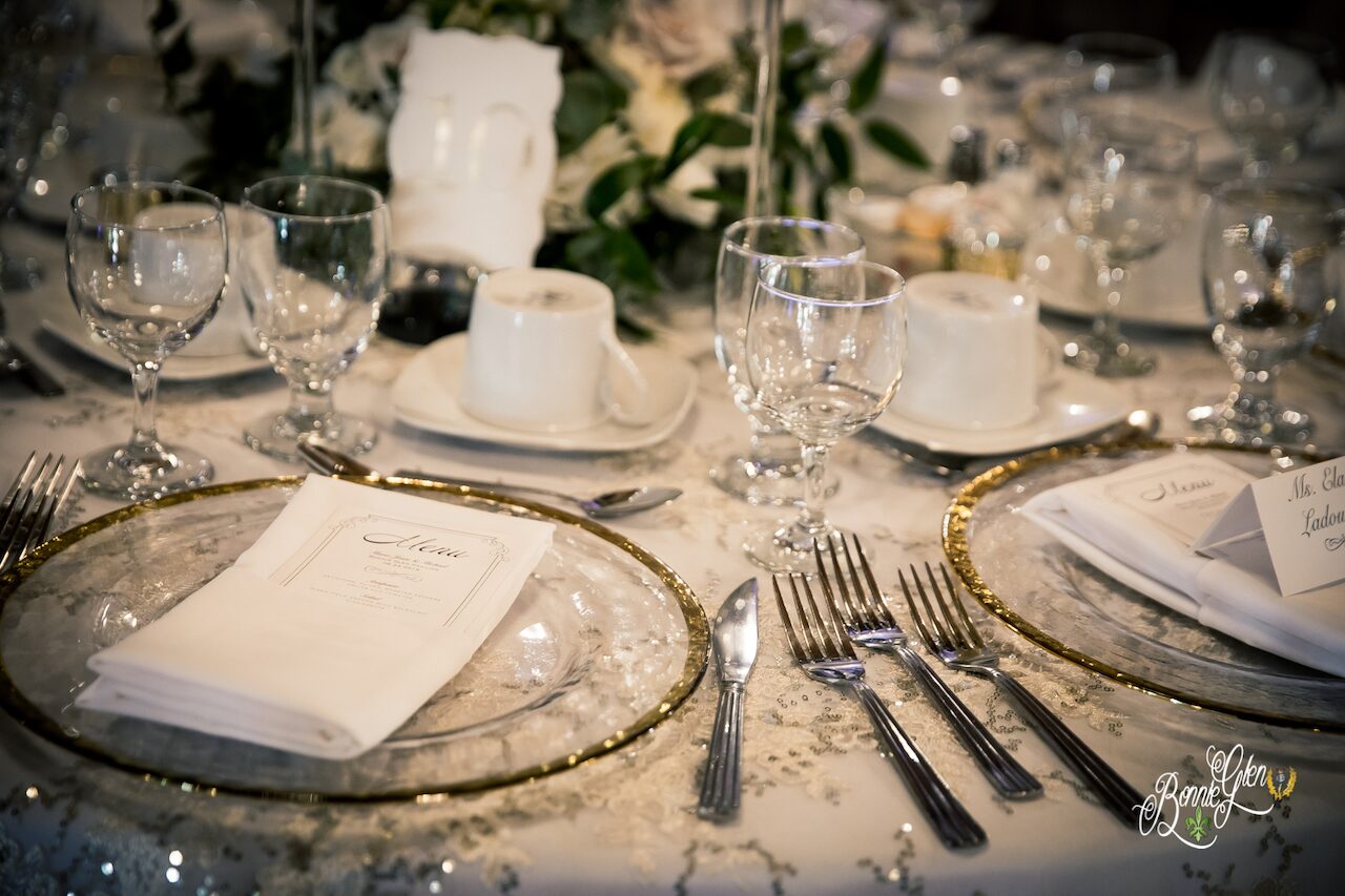 A wedding place setting at the Bonnie Glen Pavilion: gold-rimmed glass charger plates, folded napkins with printed menu cards, silverware and glasses on a lace tablecloth.